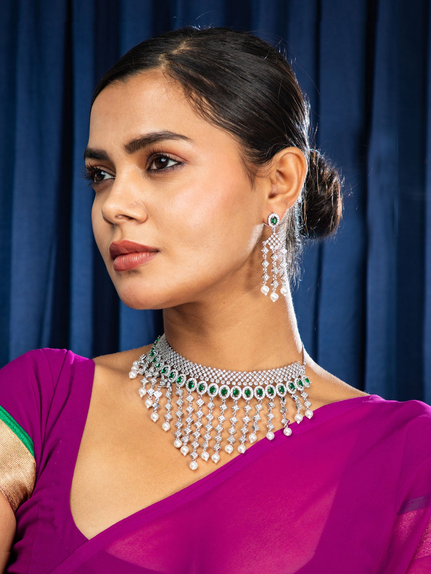 Woman wearing a purple saree with a green border and silver jewelry against a blue curtain background