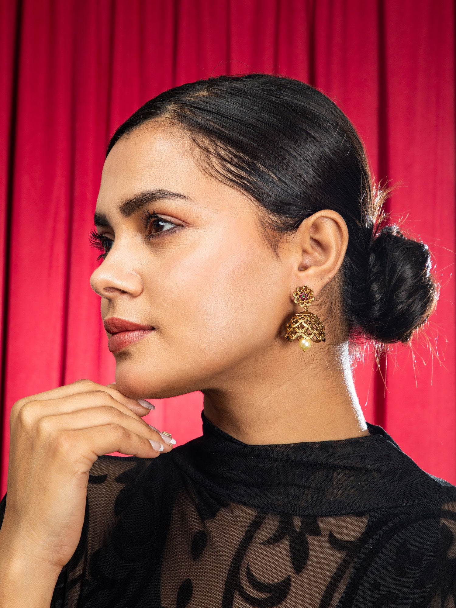 Woman with gold plated jhumka earrings​ against a red curtain background.